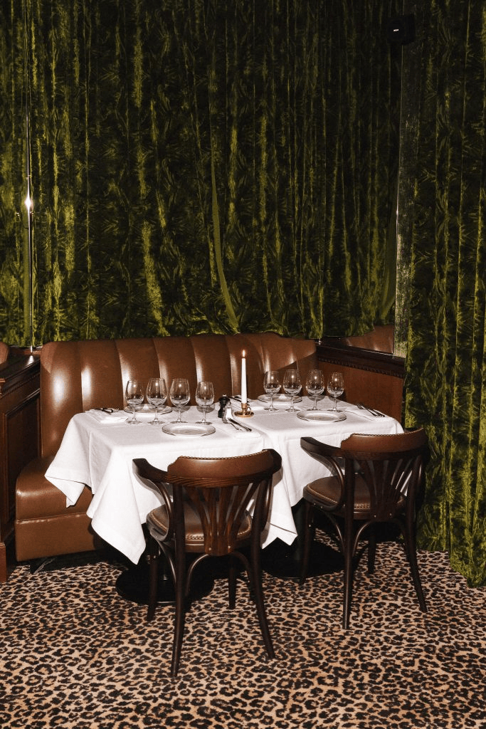 A cozy restaurant table set for dinner, featuring a white tablecloth, wine glasses, and a candle. The background is draped in green velvet curtains, and the floor has a leopard print design.