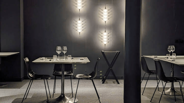 Modern restaurant interior featuring a sleek table with wine glasses, stylish black chairs, and decorative lighting on a dark wall.