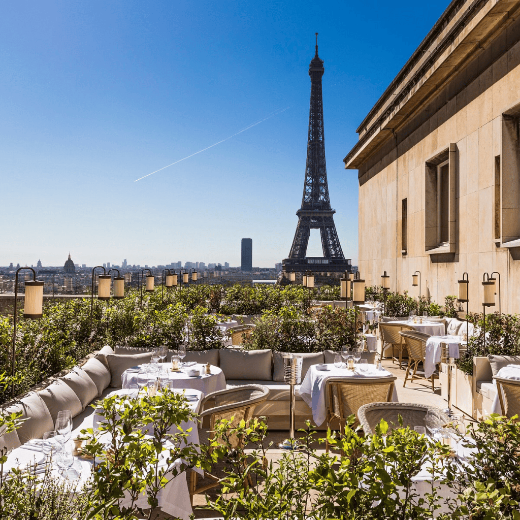 Rooftop terrace dining area with tables and chairs, surrounded by greenery, overlooking the Eiffel Tower under a clear blue sky.
