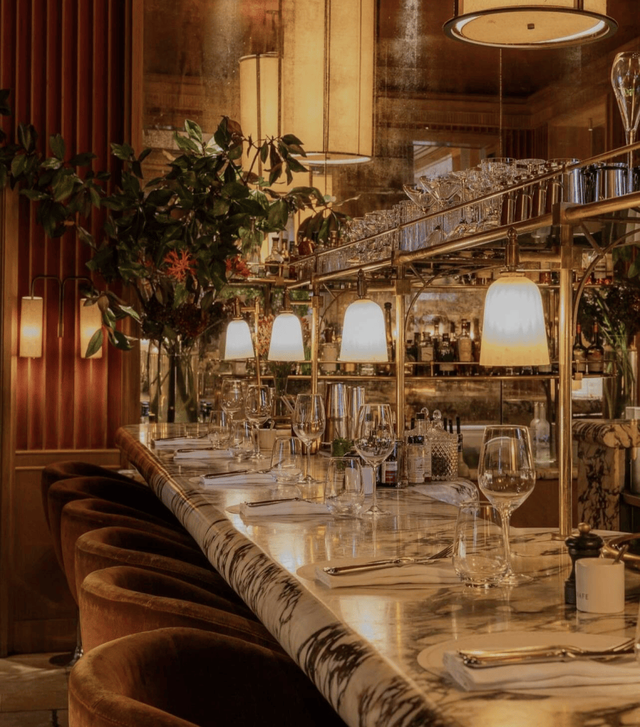Interior of a stylish restaurant featuring a marble bar, elegant glassware, and warm lighting. Upholstered seating in rich brown tones complements the decorative plants and bar shelves stocked with spirits.