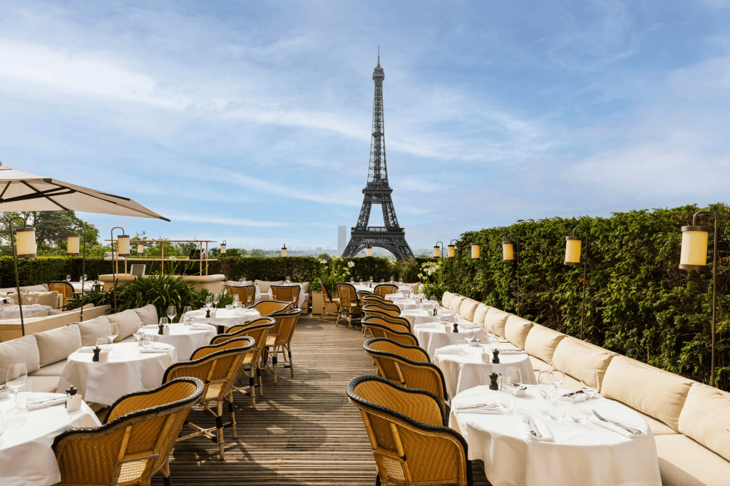 Outdoor dining area with tables set for guests, featuring wicker chairs and white tablecloths, overlooking the Eiffel Tower under a clear blue sky.