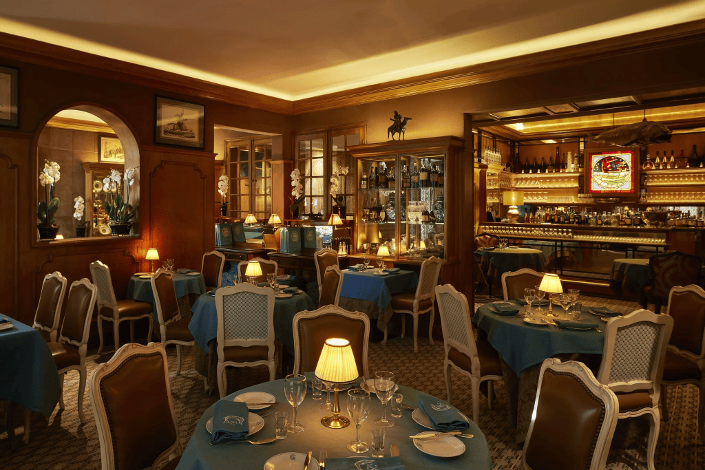 Interior view of an elegant restaurant featuring blue tablecloths, ornate chairs, and soft lighting, with a well-stocked bar and decorative elements.