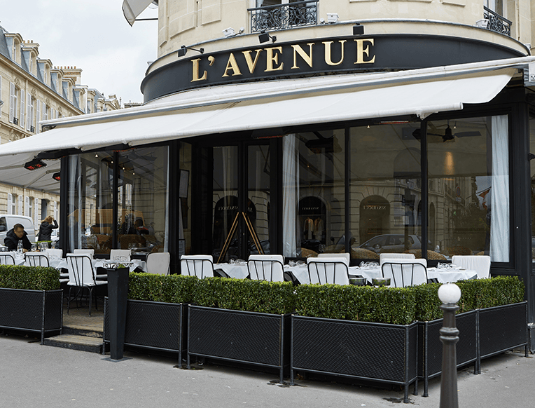 Exterior view of L'Avenue restaurant with outdoor seating and white tablecloths, surrounded by trimmed hedges.