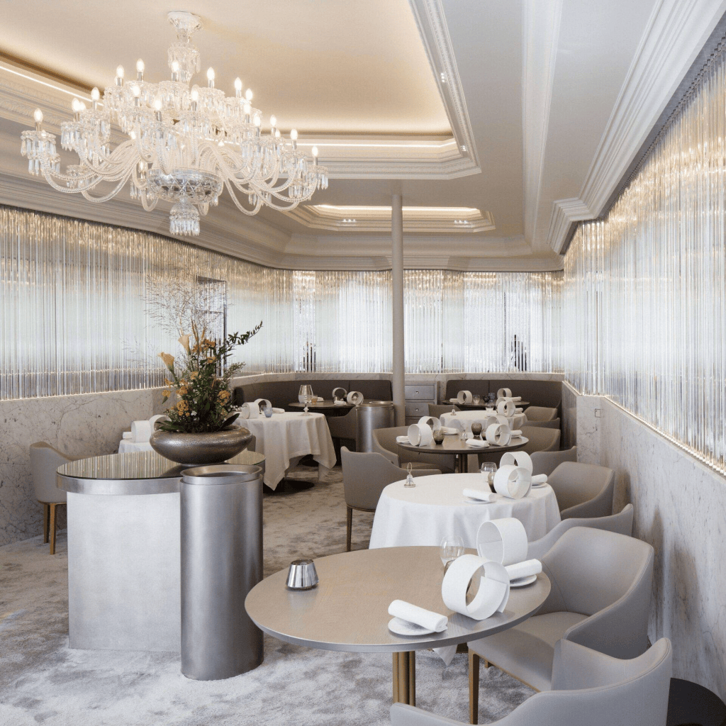 Elegant dining room with a chandelier, featuring round tables with white tablecloths and modern chairs, a floral centerpiece, and large windows covered with vertical blinds.