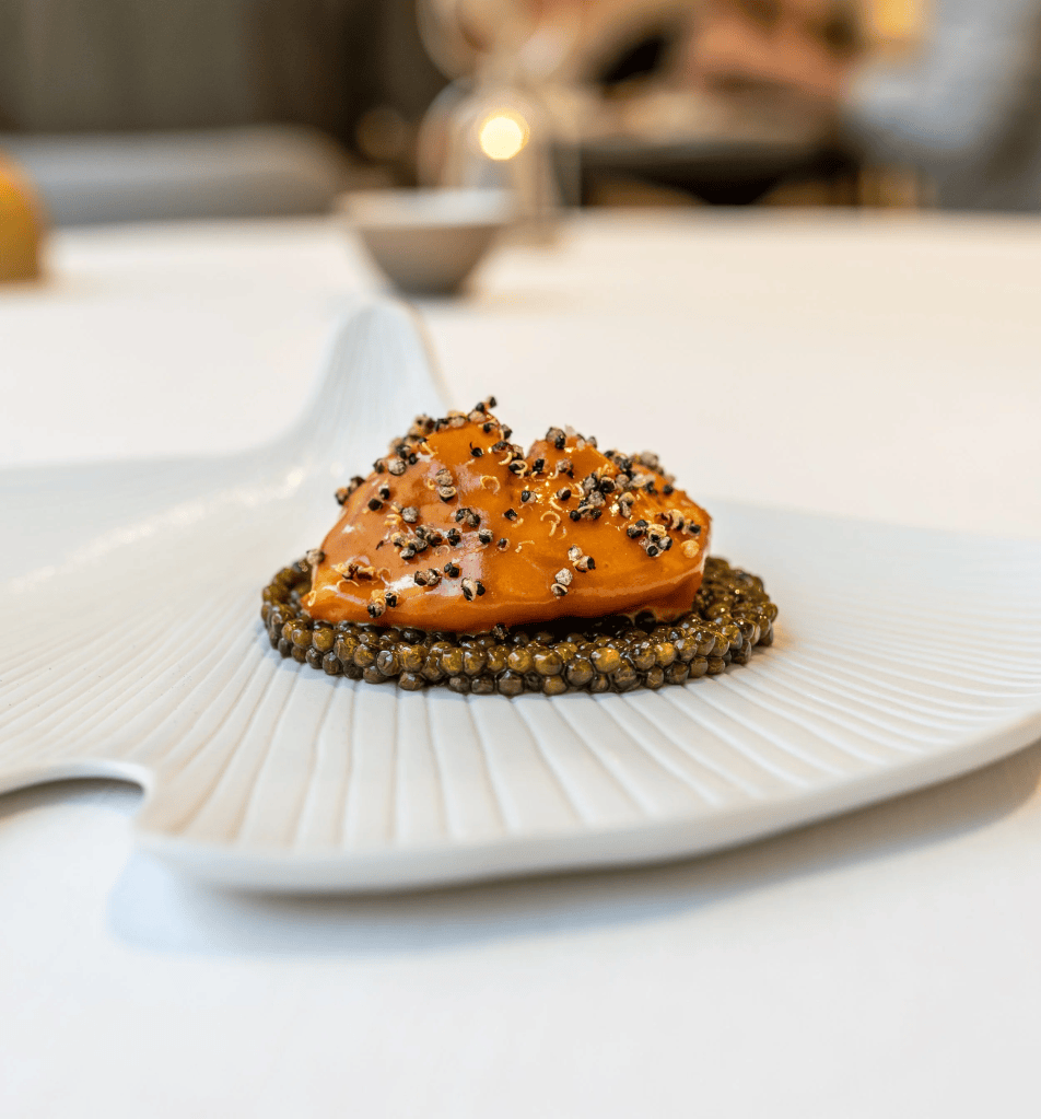 A beautifully presented gourmet dish featuring a bright orange topping sprinkled with black seeds, served on a textured white plate with a circular base of small dark beads resembling caviar.