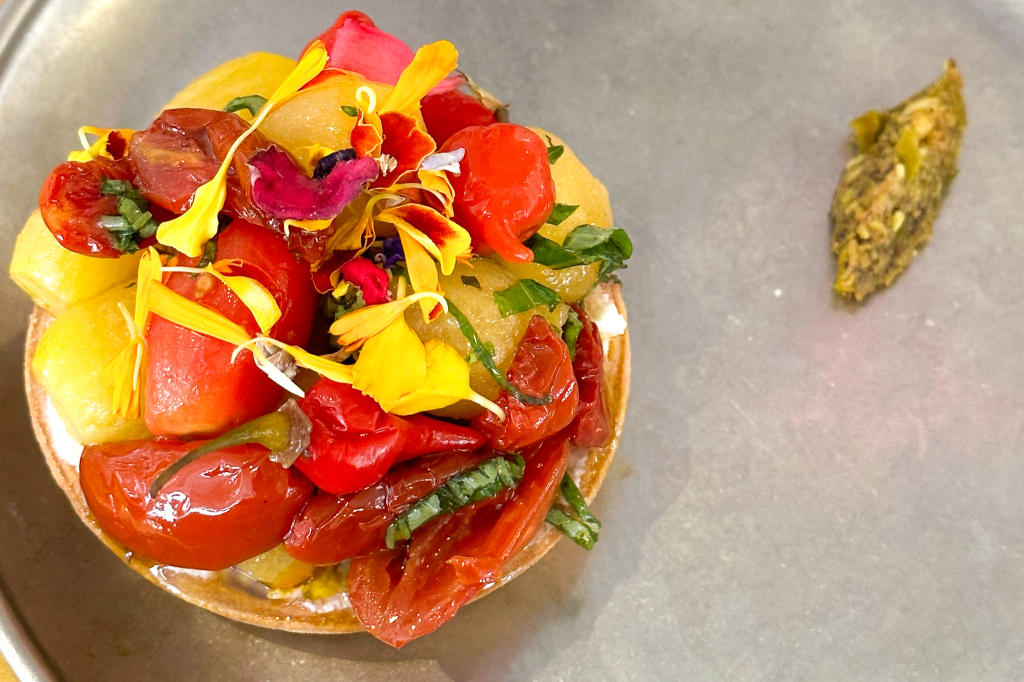 A vibrant tart topped with assorted roasted tomatoes, yellow squashes, and edible flowers, served on a metallic plate with a small herb paste on the side.