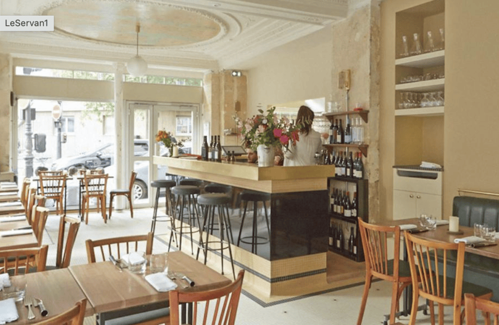 Interior of a stylish restaurant featuring wooden tables and chairs, a bar area with a gold and black design, and large windows letting in natural light.