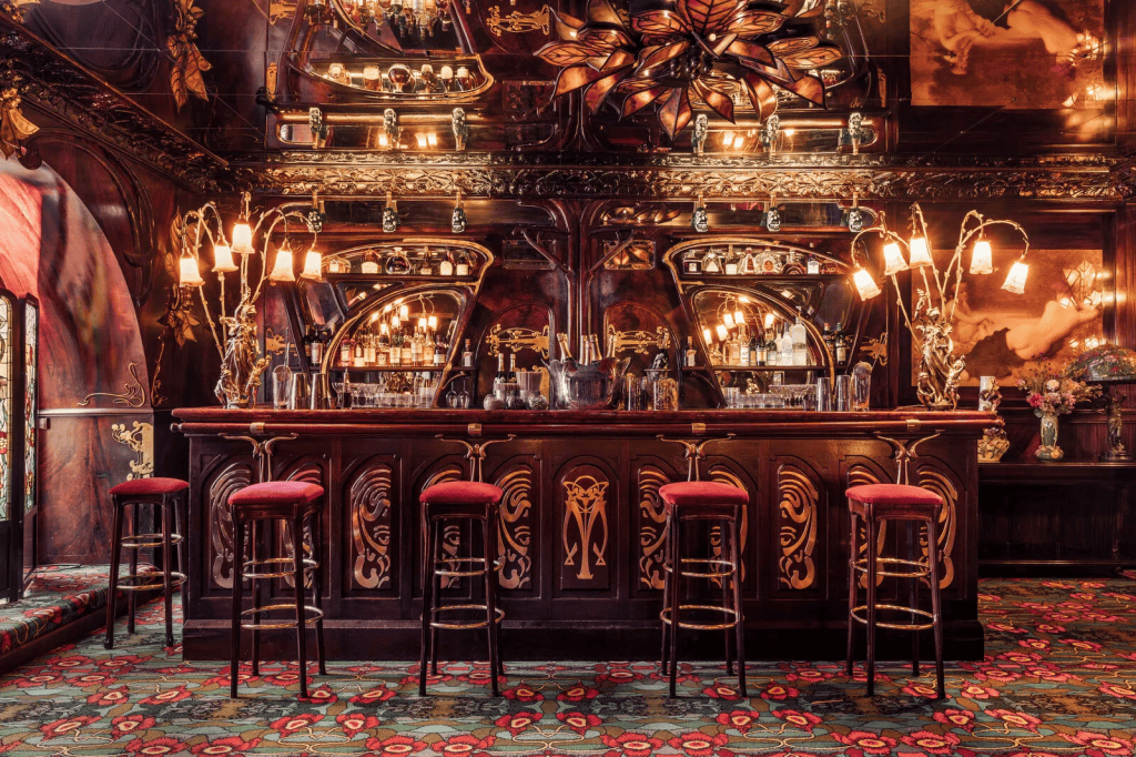 Elegant bar interior featuring ornate wooden detailing, decorative mirrors, and stylish seating with red upholstery.
