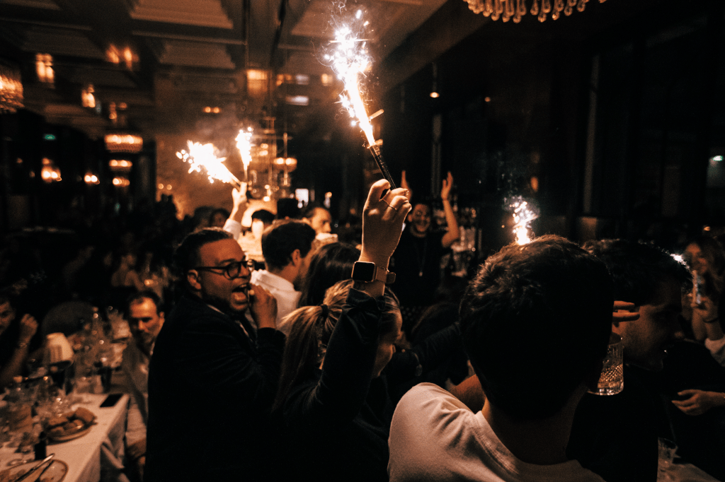A lively celebration scene in a restaurant with people raising sparklers, surrounded by a crowd enjoying dinner and drinks. Dim lighting and elegant decor create a festive atmosphere.