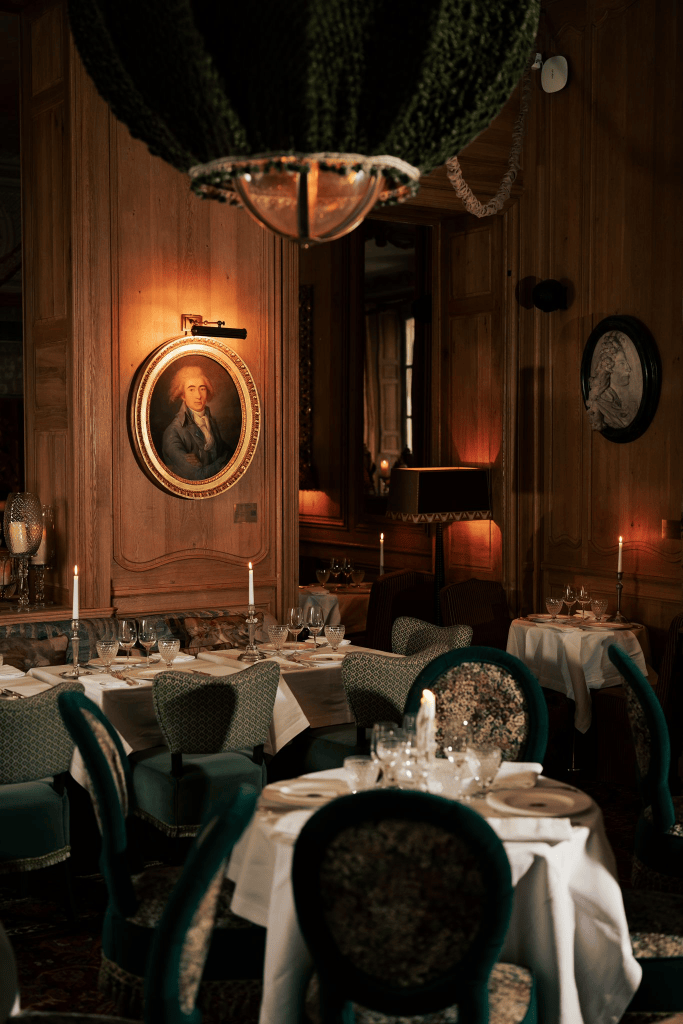 Interior of an elegant restaurant featuring neatly set tables, plush green chairs, and a chandelier, with a historical portrait on the wooden wall.