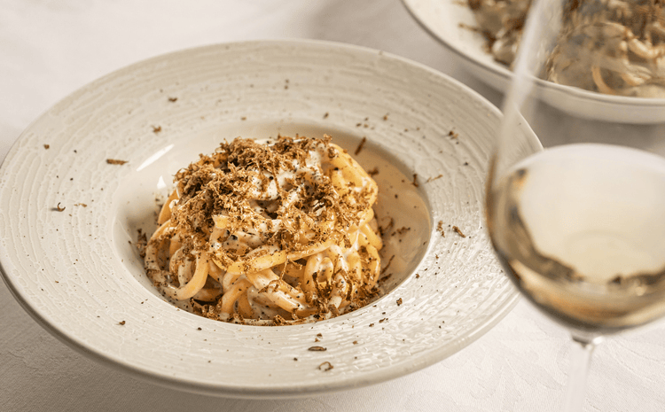 A plate of pasta topped with grated truffles, served with a glass of white wine.