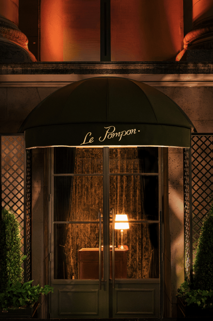Entrance of Le Pompon restaurant with a dark green awning and illuminated interior featuring a lamp and decorative curtains.