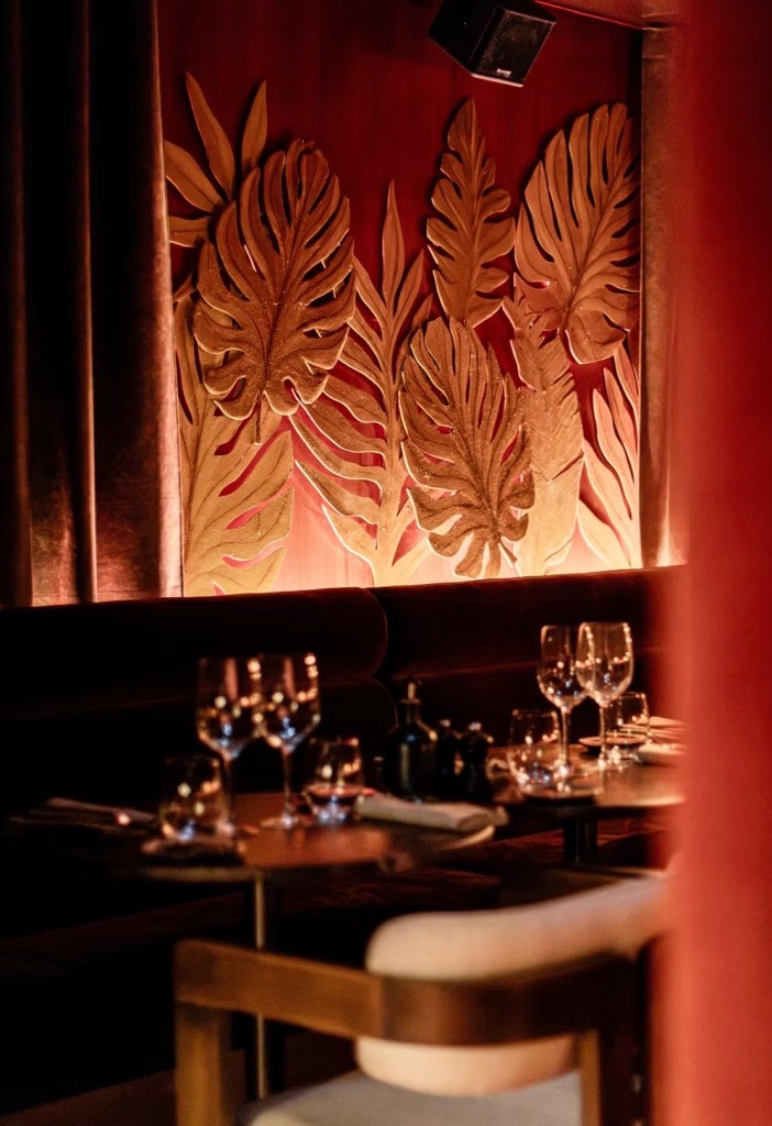 A stylish restaurant interior featuring a wall with large, golden leaf decorations illuminated by soft lighting, with elegantly set tables in the foreground.